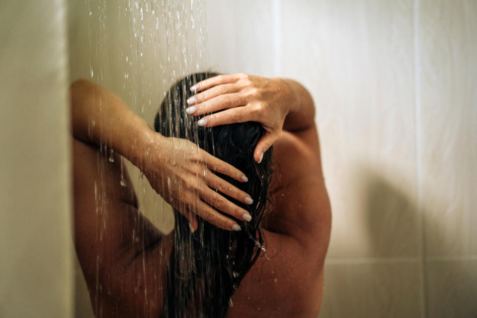 Person with back to camera stands in a shower, hands on head and water flowing down