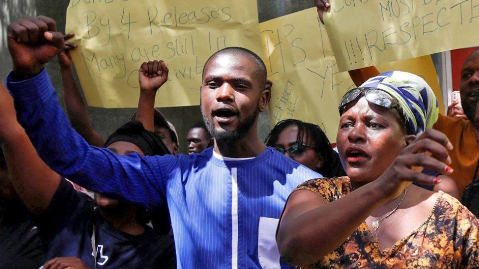 One of a group of Kenyan protesters holding up yellow handwritten posters calling for the release of people who have been kidnapped. Several—including a young bearded man in a blue-collared shirt and a woman wearing a brown patterned v-neck, necklace, blue-and-white headscarf and sunglasses on her forehead in close-up—have raised their fists.  