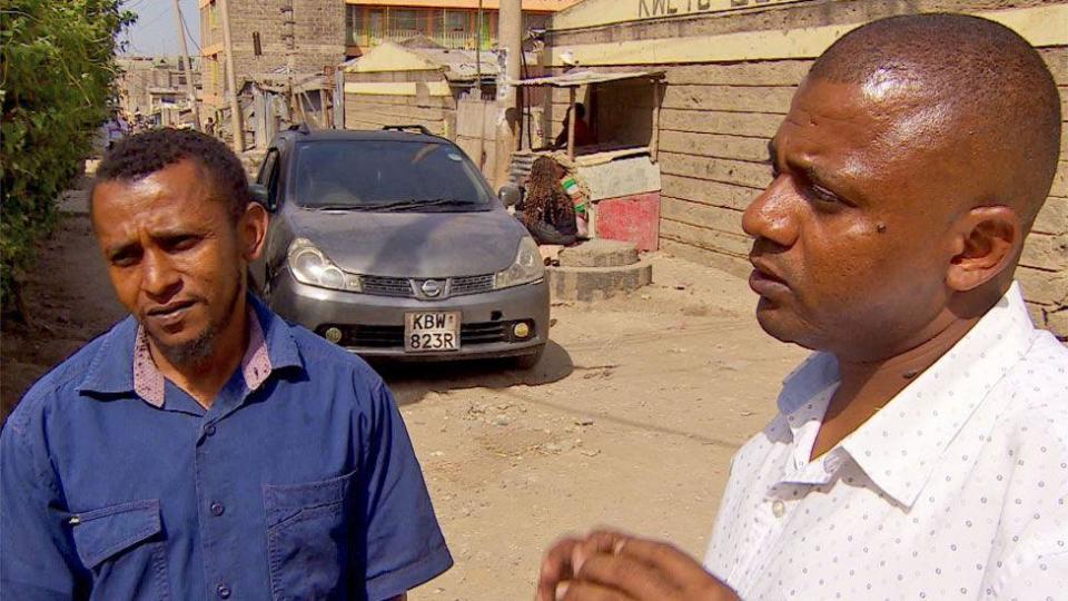 Aslam Longton in a blue shirt and Jamil Longton and a white shirt with a dot pattern talk while standing on a dirt road in Kitengela town. A silver car can be seen behind them.