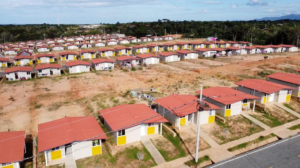 Rows of identical gray and yellow houses with red roofs along roads, with suddenly empty soil behind every house and forest -covered hills in the background, in Isberyala