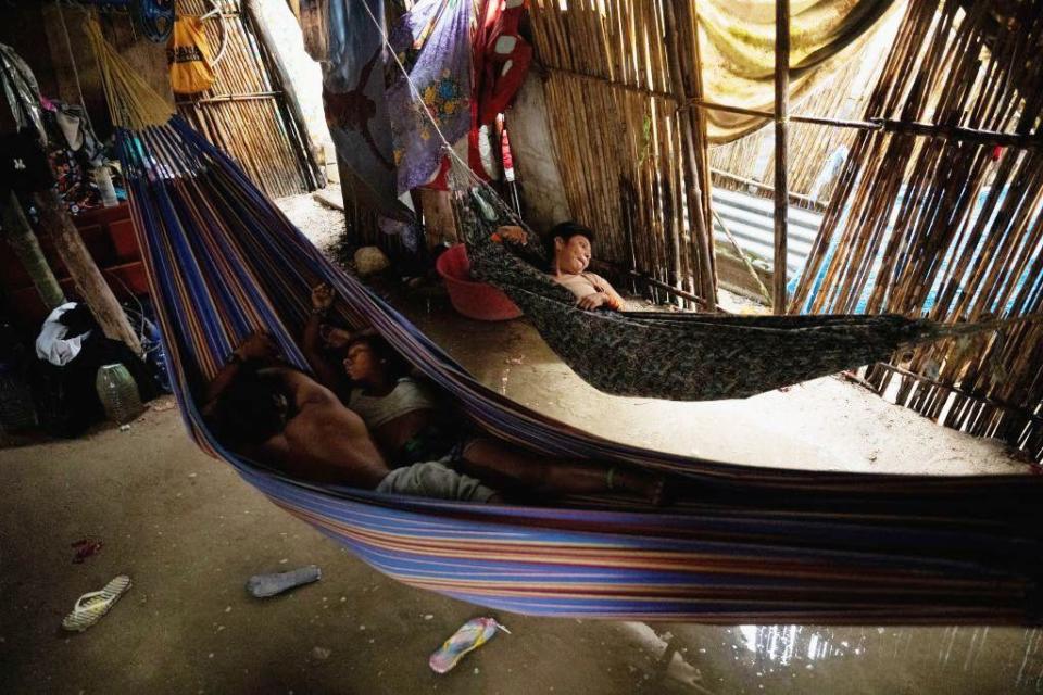 Two people are in one hammock and another in a separate hammock, built in a room from wooden posts. There is shallow water on the ground under the hammocks, with two sandals floating in the water.