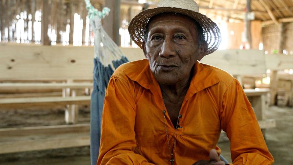 Tito López, the community leader of Isberyala, in a bright orange shirt and a straw hat, is in a hammock, looking at the camera with a thoughtful expression. Wooden benches and the wooden walls and the roof of the meeting house can be seen behind him.