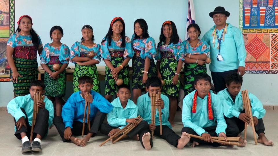 Seven girls stand behind six boys with their teacher. The boys wear turquoise shirts, the girls wear black and green molas wrapped from above the waist and turquoise flower blouses. There are brightly colored wall dresses in the background.