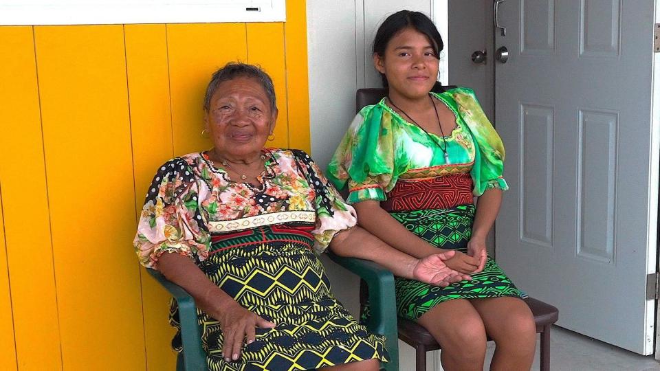 Magdalena and her granddaughter Bianca, sitting on plastic seats for their new house in Isberyala. They both wear molas with clear diamond -shaped designs on it and look straight at the camera. The yellow and gray panels of their house and a door are behind them.