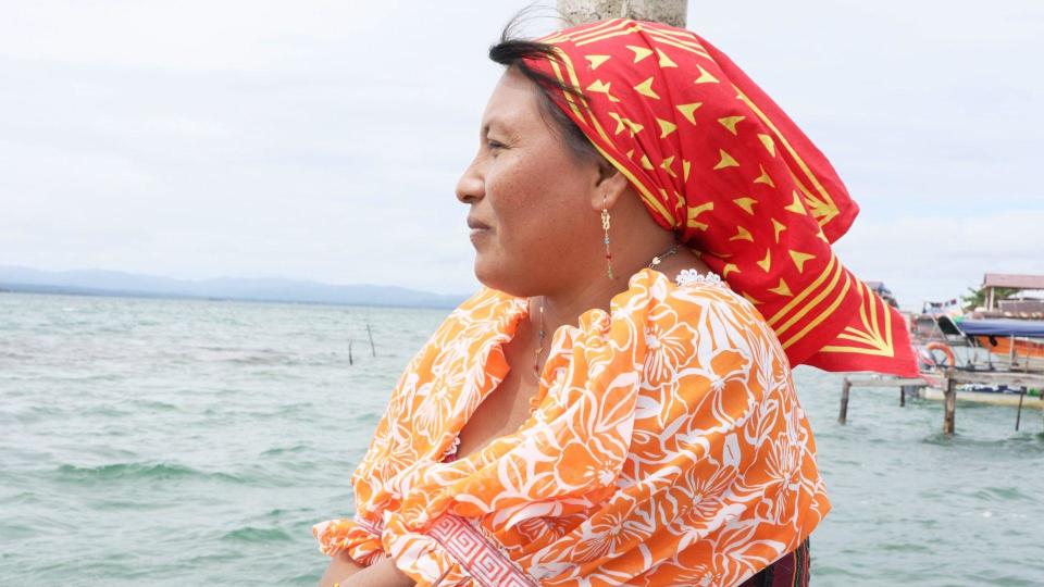 Side view of Yanisela, with a red and yellow headscarf and an orange and white flower blouse. She is looking forward to the sea with some scaffolding and buildings on the island behind her.