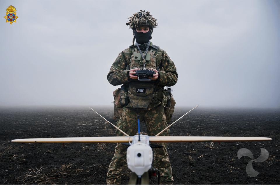 A Ukrainian drone operator who wears camouflage holds a drone controller with a fixed wingrone that flies for him. It is on black dirt with a gray background.