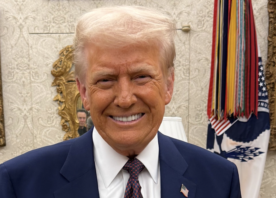 A person with stylized hair and a blue suit, with an American flag pen, laughs indoors with a decorative background