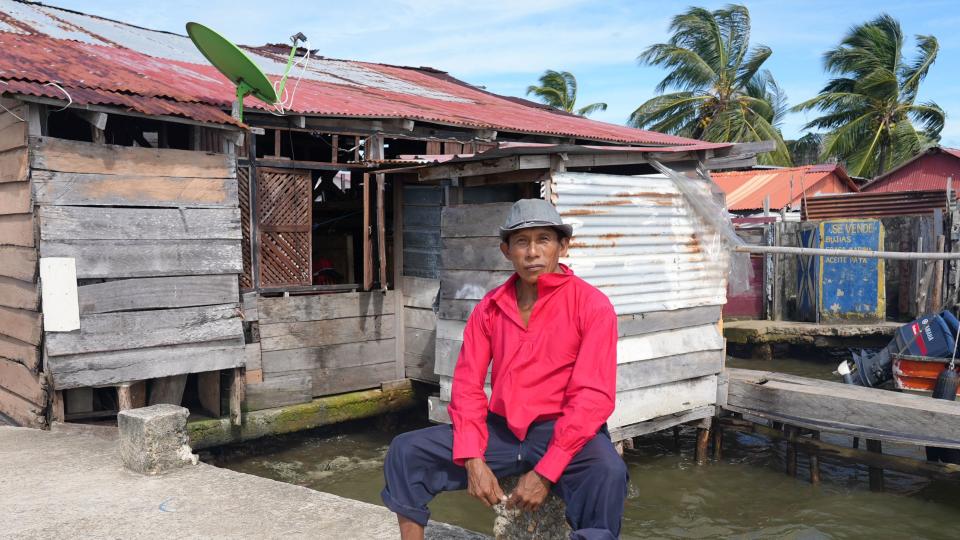 Delfino, in a bright pink shirt and gray hat, sits on a low concrete pole on a jetty, with a house built of wood and corrugated metal behind him. A part of the house is on poles and protrudes in the water.