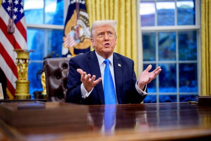 A person in a suit is at a desk with raised hands, in a formal office environment with large windows and flags visible in the background