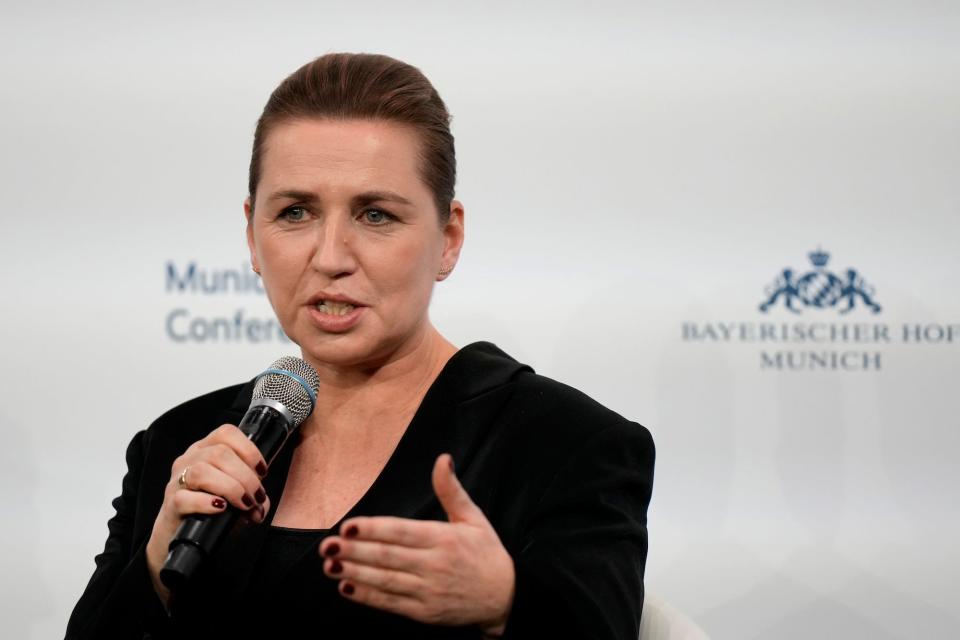 A woman with dark, pulled hair and wearing a dark jacket speaks in a microphone for a white background with a blue hotellogo on it