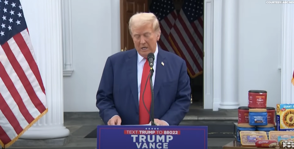 Person who speaks on a stage with text "Trump Vance" And "SMS Trump to 88022," Surrounded by flag images and displayed products