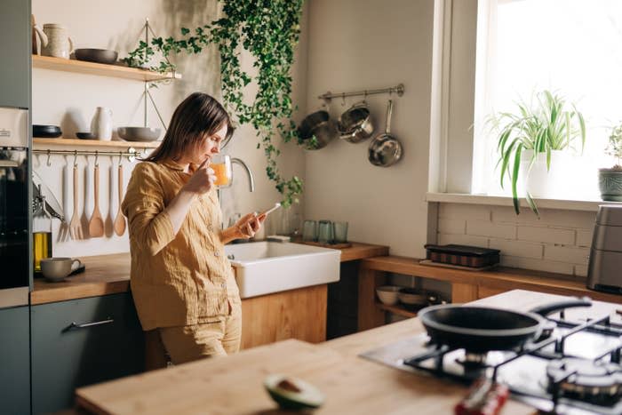 Person in a cozy kitchen, wearing comfortable, informal clothing, narrowly a drink and looks at a phone. Plants and kitchen utensils adorn the space