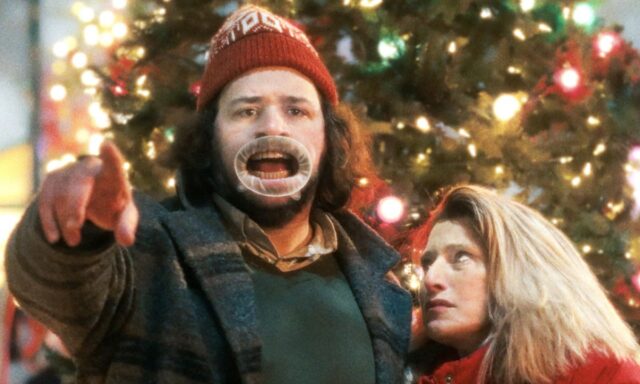A man wearing a funny hat in front of the Christmas tree robs for the camera while a woman watches and rolls her eyes