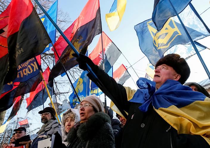 Activists in coats and hats, one draped in a flag, hold up placards and several flags.