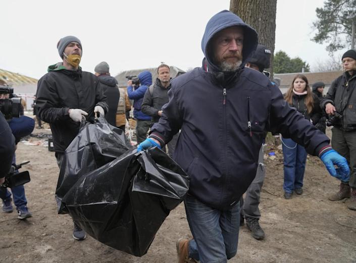 A man, right, wears a hooded jacket and blue gloves and carries a black body bag with the help of another man