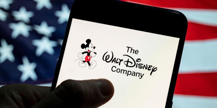 American flag in the background of someone holding a phone with the text The Walt Disney Company on the screen.