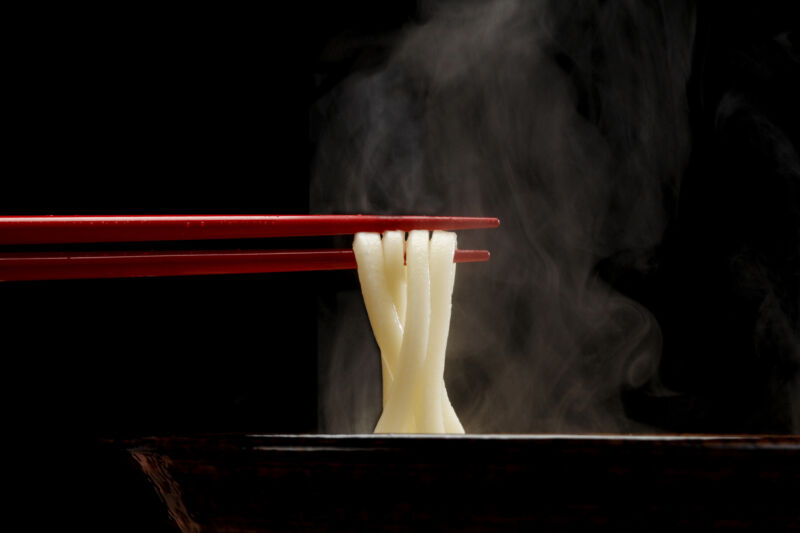 Japanese Sanuki Udon, thick straight noodles, lifted from soup bowl with red chopsticks.  Steam looks sharp against a black background.  Angle at eye level.