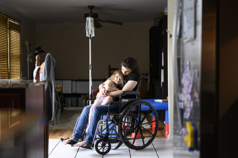 A tall COVID patient sits with her daughter in her wheelchair as she receives a saline drip at her home in Maryland on Friday, May 27, 2022.  