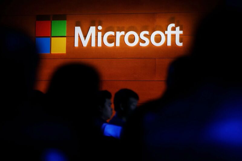 Shadowy figures stand below a Microsoft logo on a faux wood wall.