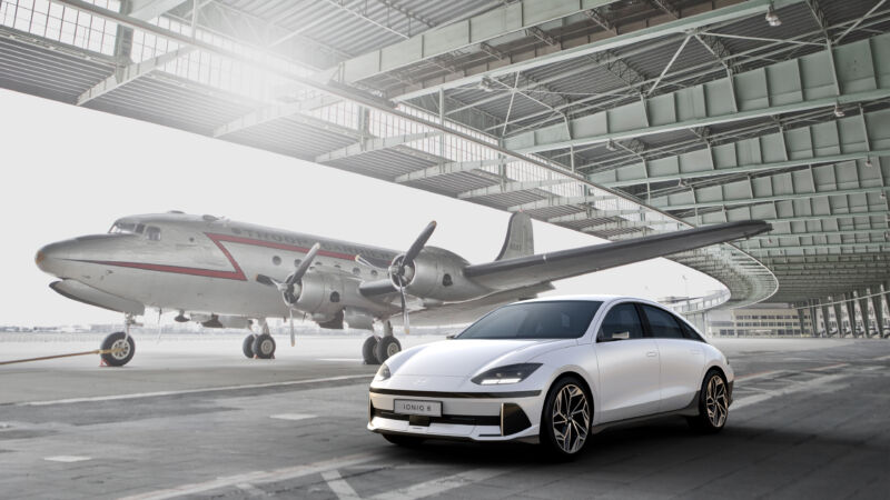 A white Hyundai Ioniq 6 sedan in front of a 1930s passenger plane in a hangar.