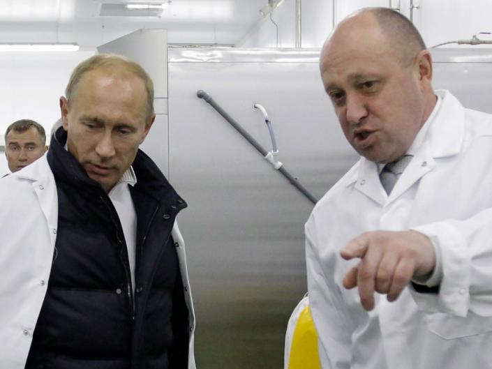 Putin and Prigozhin in white coats, with the latter pointing to something off-camera