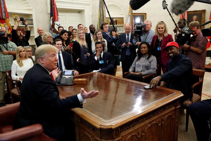 Donald Trump, behind his desk in the Oval Office, with a throng of cameramen and visitors surrounding him, makes a joke that most of the assembled party find highly amusing.  Seated are Ivanka Trump and Jared Kushner, as well as Kanye West, wearing a red MAGA baseball cap.