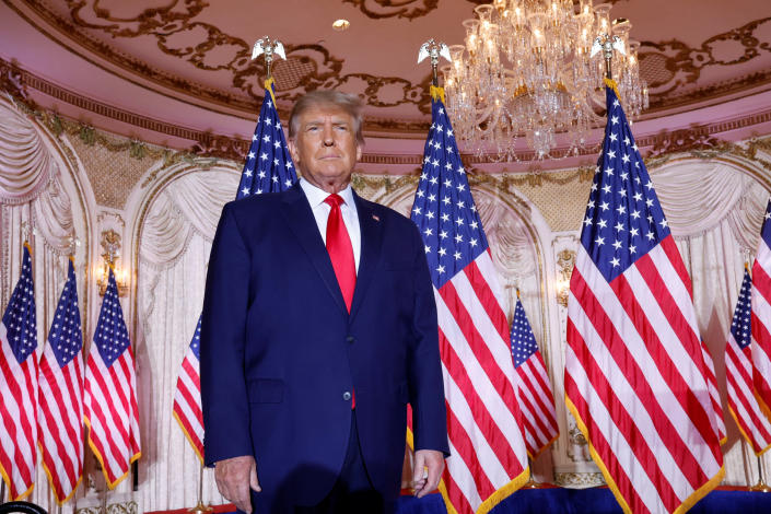 Donald Trump, hands by his side under a chandelier in the Mar-a-Lago ballroom, with eight American flags visible behind him.