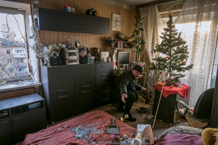 A local resident, kneeling by his Christmas tree in his living room, removes shards of glass from broken windows in his apartment in a residential building.