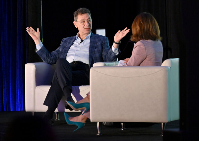 A man in a suit with an open collar addresses a woman in a matching chair.