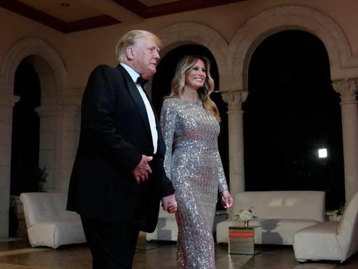 Former President Donald Trump in a black suit and bow tie holding hands with former First Lady Melania Trump in a silver dress