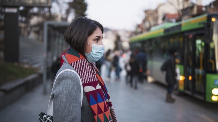 a woman is waiting for a bus wearing a medical mask