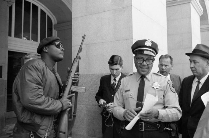 Lieutenant Ernest Holloway reads a document while a member of the Black Panther Party in a beret stands at attention, his rifle pointed in the air.