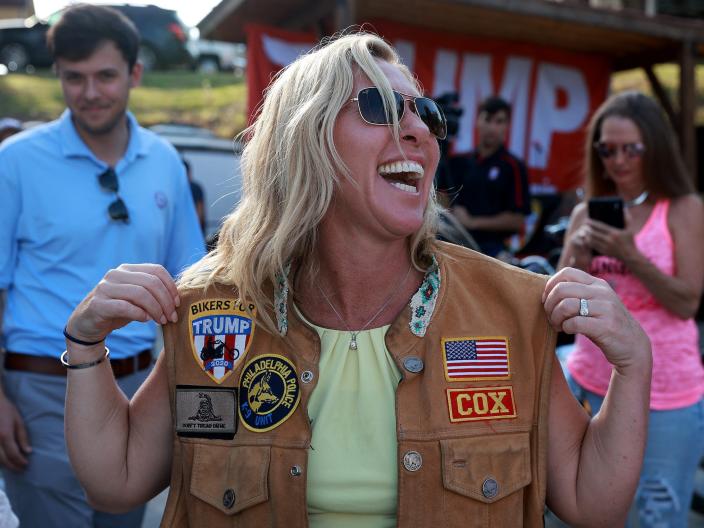 Marjorie Taylor Greene wears a motorcycle vest in front of a big Trump sign.