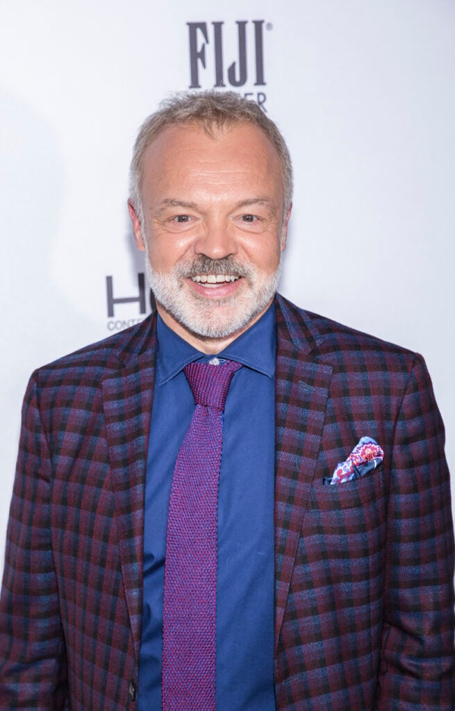 Graham Norton stands in front of a backdrop in a blue and purple blazer