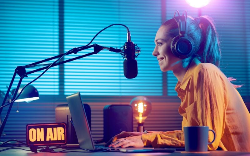 Radio presenter with ponytail and yellow shirt looking at computer and smiling while on air in a room with closed blinds