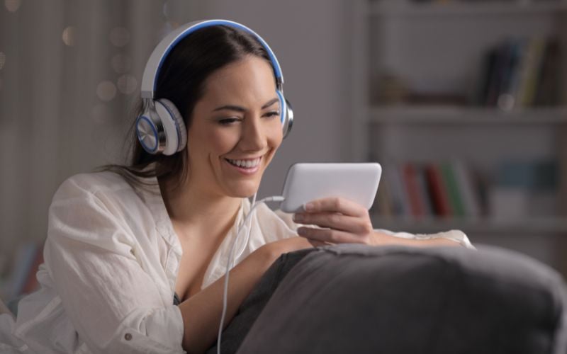 Smiling woman wearing a white shirt and headphones is looking at her phone and listening to music while sitting on her couch