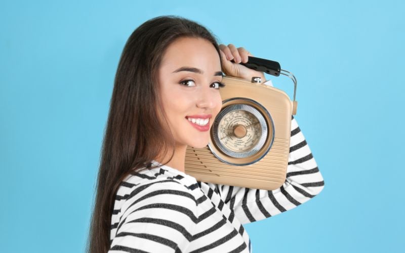 Woman in a black and white striped shirt listening to a beige DAB radio on her shoulder and smiling against a blue background in the USA