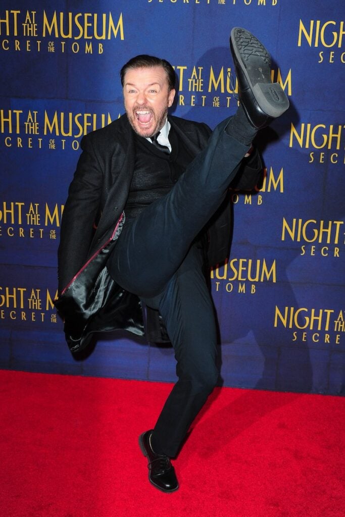 Ricky Gervais does a high leg kick on a red carpet catwalk in a black tuxedo