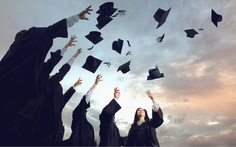 Students in graduation gowns toss graduation hats into a sunset sky