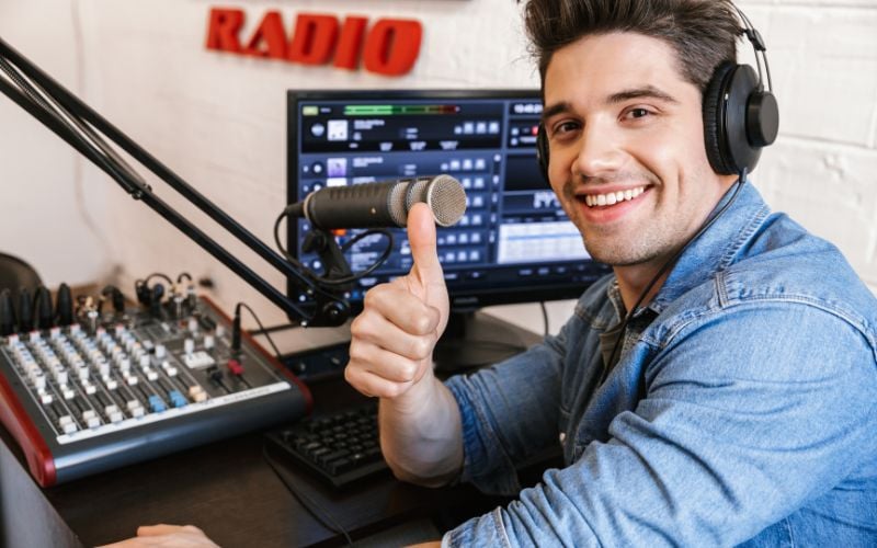 Radio host wearing a blue button down shirt with brown hair and a professional microphone in a radio studio giving a thumbs up