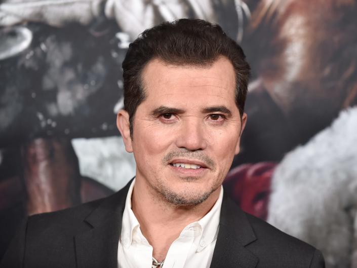 John Leguizamo poses for photos in a black suit jacket and white shirt.