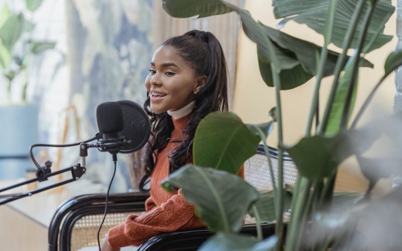 Radio announcer at the microphone in a bright room with plants in the foreground 