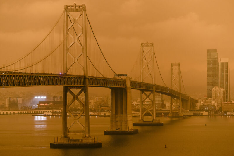 Image of an orange sky with a bridge and buildings partially disappearing into the mist.