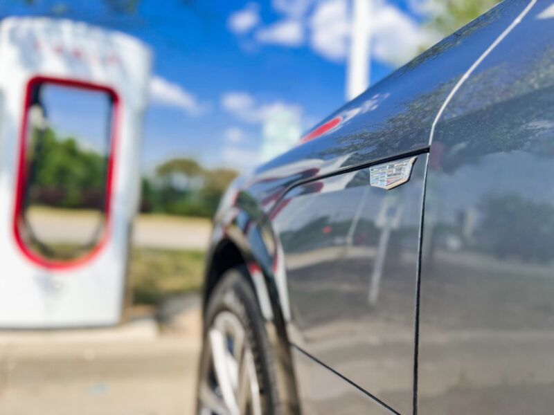 A close-up of the badge on the front fender and tailgate of a Cadillac LYRIQ parked at a Tesla Supercharger station.