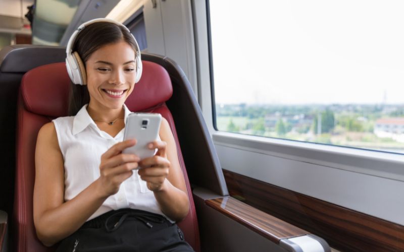 Smiling woman in white sleeveless blouse and headphones smiles while listening to a 30 second radio advertisement on her mobile phone on a train