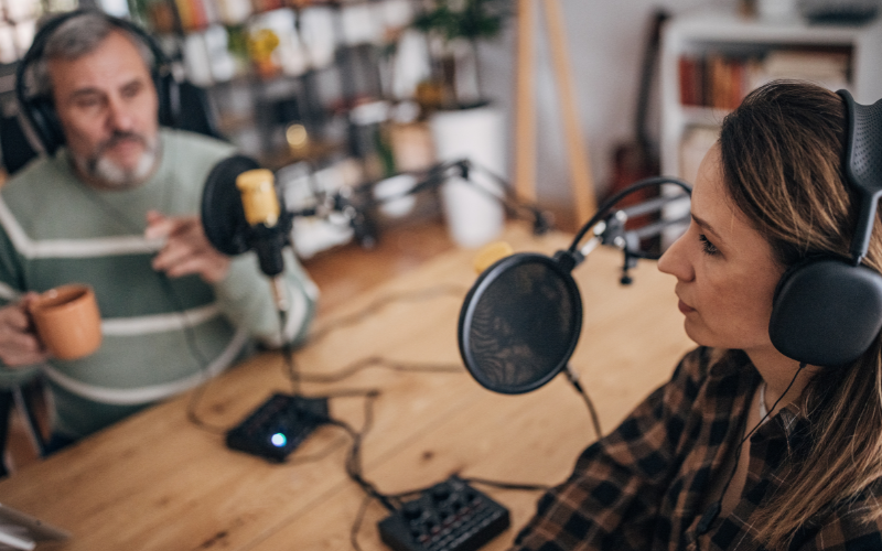 A woman is speaking to a radio host who is speaking into a microphone while a male radio host with coffee can be seen in the background