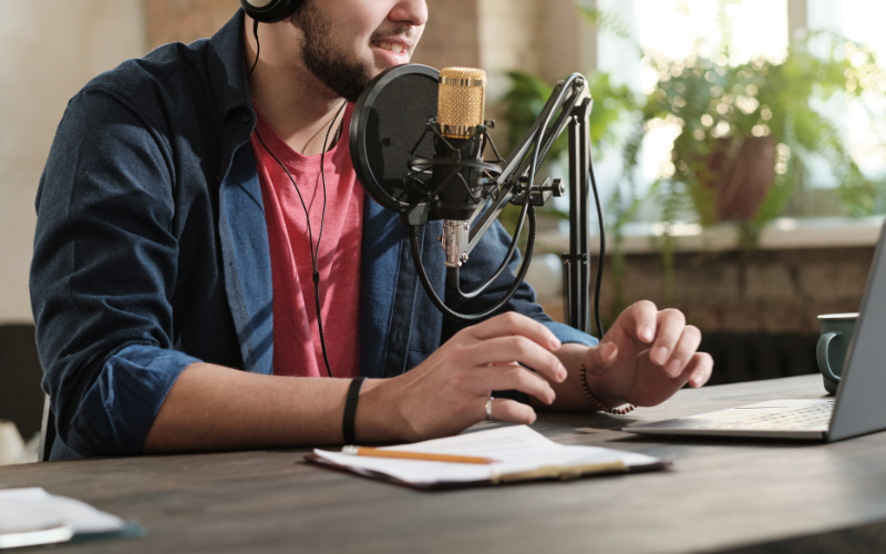 Man in a red shirt and jacket speaks into a microphone in a room with a bright window and plants