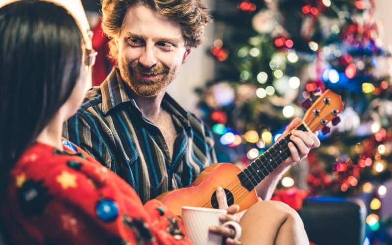 A man plays popular Christmas carols on the guitar and smiles at a woman with a Christmas tree in the background 