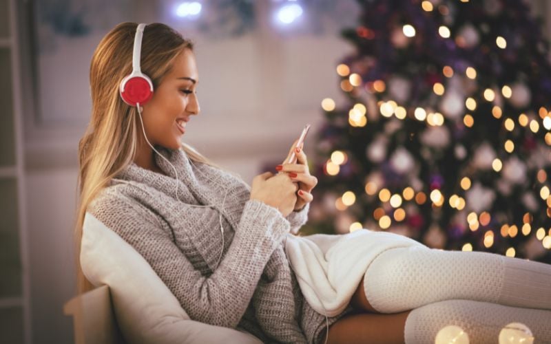 Woman on the couch, wearing a sweater and headphones, listening to music and smiling with a Christmas tree in the background