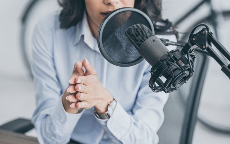 Woman from the nose down wearing a light-colored blouse, hands clasped, speaking into a microphone on Christian radio stations